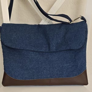 May include: A blue denim crossbody bag with a brown leather bottom panel. The bag has a flap closure and a navy blue adjustable strap. The bag is sitting on a white chair.