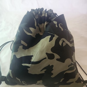 May include: A drawstring backpack with a camouflage pattern in shades of green, black, and tan. The bag is gathered at the top with a black drawstring closure. The background is plain white.