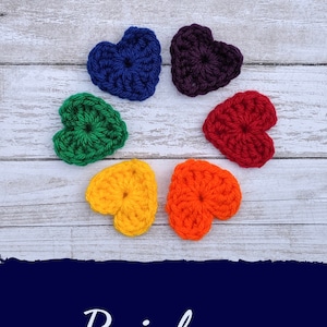 May include: Six crocheted hearts in rainbow colors, arranged in a circle on a white wooden surface. The colors are red, orange, yellow, green, blue, and purple.
