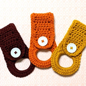May include: Three crocheted dishcloths in orange, brown, and yellow with white buttons. The dishcloths are made with a simple stitch pattern and have a loop for hanging.