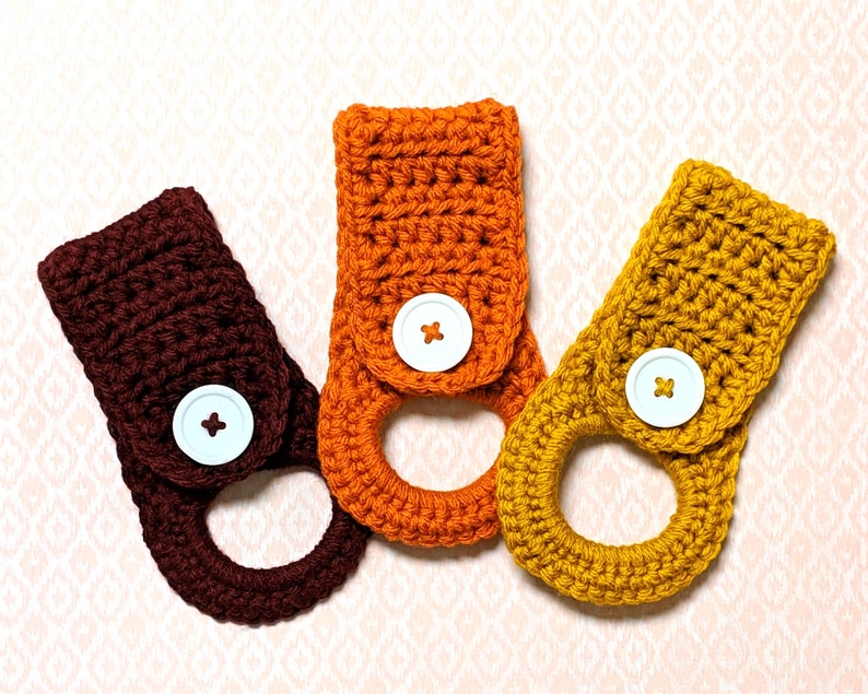May include: Three crocheted dishcloths in orange, brown, and yellow with white buttons. The dishcloths are made with a simple stitch pattern and have a loop for hanging.