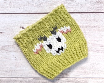 Op de afbeelding: Een groene gebreide koffiewarmer met een wit, zwart en grijs geborduurd gezicht. Het gezicht heeft twee zwarte ogen, een witte neus en een zwarte mond.