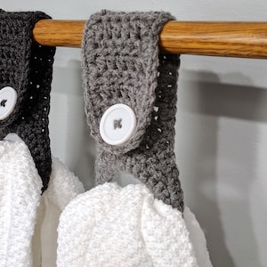 May include: Two crocheted towel holders with white buttons hanging on a wooden rod. The towel holders are gray and black.