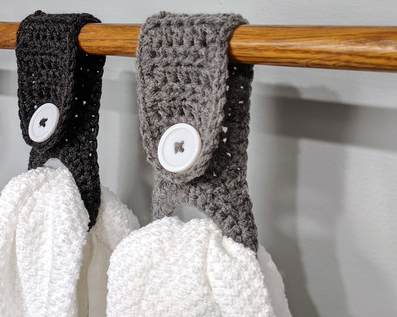 May include: Two crocheted towel holders with white buttons hanging on a wooden rod. The towel holders are gray and black.