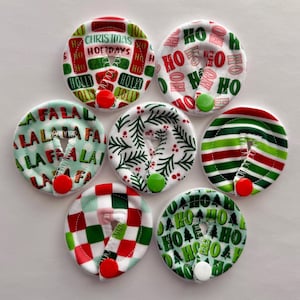 May include: Set of six Christmas-themed fabric appliques with various designs, including "Ho Ho Ho", "Fa La La", and a green and red striped pattern. Each applique has a white button in the center.