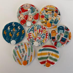 May include: Seven circular fabric pads with various school-themed patterns including apples, yellow school buses, alphabet letters, backpacks, pencils, and colorful stripes. Each pad features a small white or colored plastic snap closure.