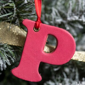 May include: A bright pink leather ornament shaped like the letter "P" hanging from a red ribbon on a snowy Christmas tree.
