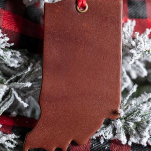 May include: A brown leather ornament shaped like the state of Indiana, hanging from a red ribbon on a snowy Christmas tree.