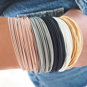 K&ouml;nnte beinhalten: Ein Stapel d&uuml;nner Metallarmb&auml;nder in Ros&eacute;gold, Silber, Schwarz und Gold. Die Armb&auml;nder sind &uuml;bereinander gelegt und erzeugen so einen einzigartigen und stilvollen Look.
