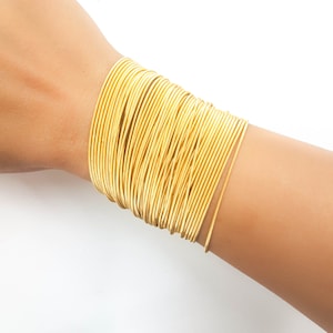 Peut inclure: Un bracelet doré composé de multiples bandes fines en métal empilées les unes sur les autres. Le bracelet est porté au poignet.