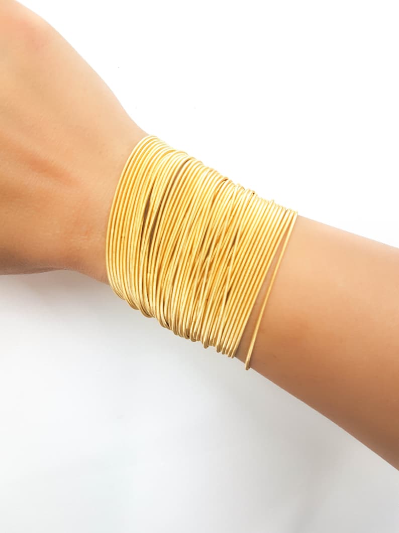 May include: A gold-toned bracelet made of multiple thin, stacked metal bands. The bracelet is worn on a person's wrist.