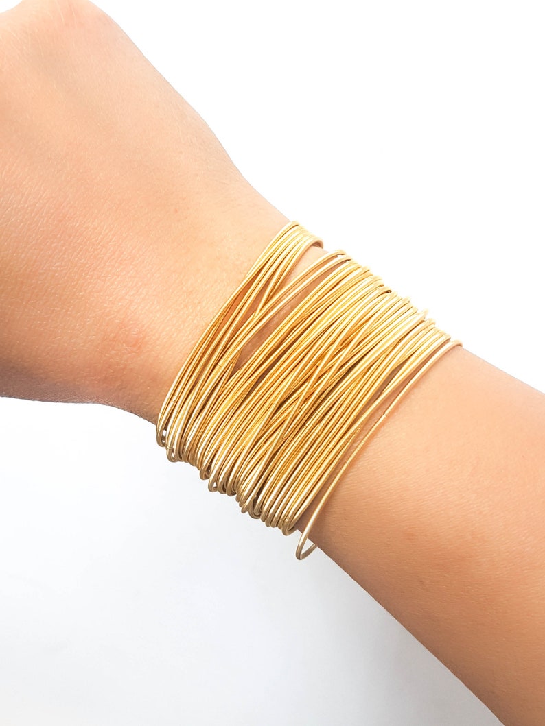 K&ouml;nnte beinhalten: Ein goldfarbenes Armband aus mehreren d&uuml;nnen, miteinander verwobenen Metallb&auml;ndern.
