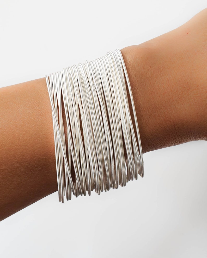 May include: A stack of silver bangle bracelets on a person's wrist. The bracelets are thin and delicate, and they are arranged in a neat and orderly fashion.