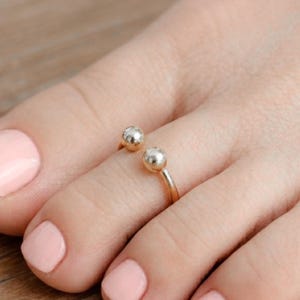 Könnte beinhalten: Ein goldfarbener Zehenring mit zwei kleinen, silberfarbenen Kugeln. Der Ring ist offen, so dass er an verschiedene Zehengrößen angepasst werden kann. Die Kugeln sind glatt und glänzend und verleihen dem schlichten Design einen Hauch von Eleganz.
