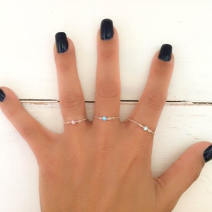 Peut inclure: Une main avec des ongles noirs vernis porte trois anneaux délicats avec de petites pierres précieuses. Les anneaux sont en argent et présentent une pierre précieuse rose, bleue et blanche.