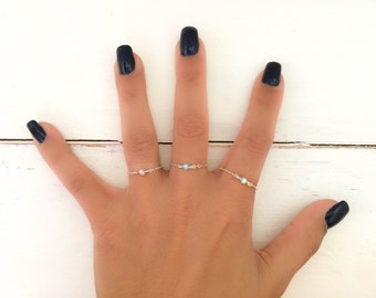 May include: A hand with black nail polish is wearing three delicate rings with small gemstones. The rings are silver and feature a pink, blue, and white gemstone.