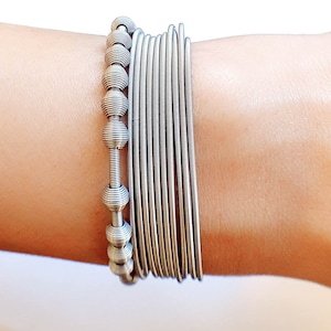 Könnte beinhalten: Ein silbernes Armband mit mehreren dünnen, gewickelten Metallbändern und einem Perlenanhänger mit einem strukturierten, abgerundeten Design.