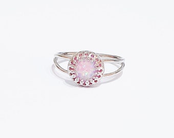 May include: A silver ring with a round, light pink opal stone. The opal is surrounded by a decorative, scalloped setting. The ring has a split shank design, with two bands that curve around the finger.