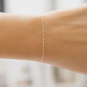 May include: A delicate silver chain bracelet. The bracelet is composed of small, oval links. The bracelet is shown on a person's wrist, highlighting its simple and elegant design. The bracelet is a minimalist piece of jewelry.