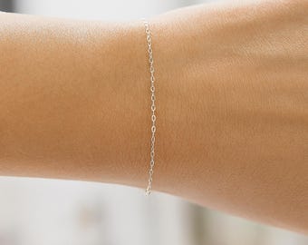 May include: A delicate silver chain bracelet. The bracelet is composed of small, oval links. The bracelet is shown on a person's wrist, highlighting its simple and elegant design. The bracelet is a minimalist piece of jewelry.