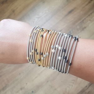 Pode incluir: Um conjunto de pulseiras elásticas douradas e prateadas com contas pretas. As pulseiras são usadas no pulso.