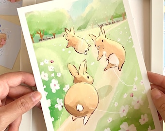 Puede incluir: Una impresión artística enmarcada con tres conejos de dibujos animados en un campo de flores blancas. La impresión está sostenida por manos y rodeada de otras tarjetas pequeñas con ilustraciones de animales. La obra de arte tiene una paleta de colores pastel.