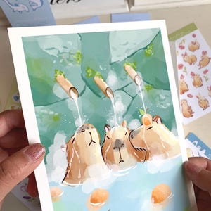 May include: A watercolour print showing three capybaras in a hot spring. The artwork depicts the animals with tan fur, submerged in blue water, with water flowing from bamboo spouts. Orange fruit floats in the water.