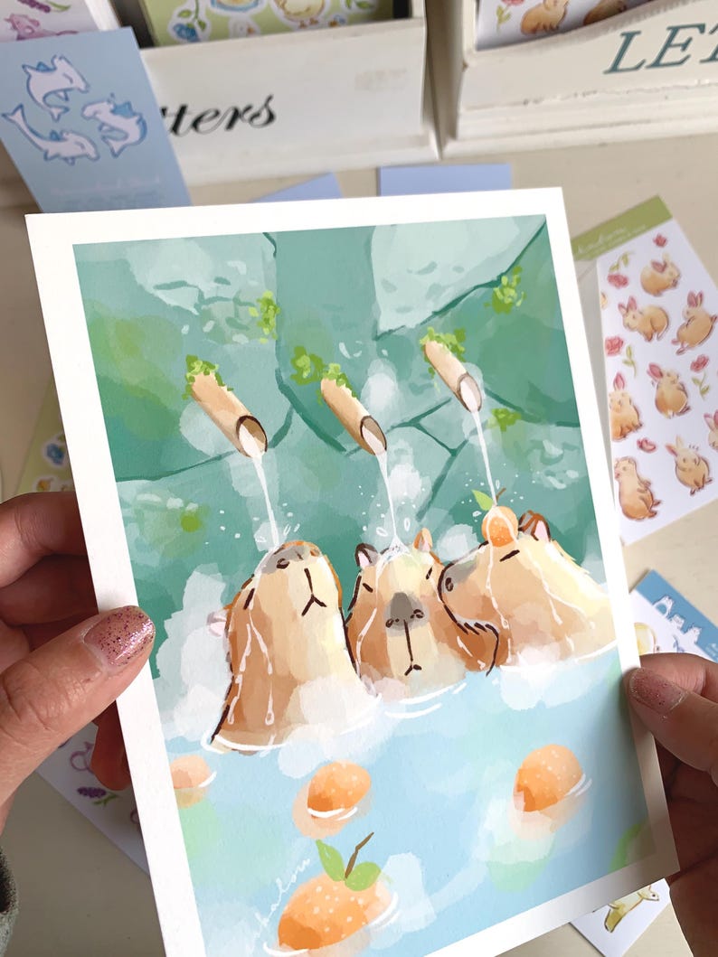 May include: A watercolour print showing three capybaras in a hot spring. The artwork depicts the animals with tan fur, submerged in blue water, with water flowing from bamboo spouts. Orange fruit floats in the water.