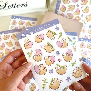 May include: A sheet of stickers featuring cute sloth illustrations. The sloths are depicted in various poses, including hanging from branches, holding hearts, and sleeping. The stickers are printed on white paper with a glossy finish. The sheet is labeled "eshalom" and "cute prints, cards, stickers & more".
