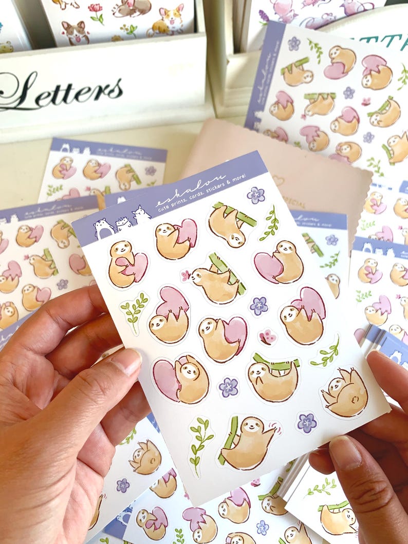 May include: A sheet of stickers featuring cute sloth illustrations. The sloths are depicted in various poses, including hanging from branches, holding hearts, and sleeping. The stickers are printed on white paper with a glossy finish. The sheet is labeled "eshalom" and "cute prints, cards, stickers & more".