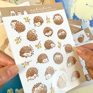 May include: A sheet of stickers featuring various illustrations of hedgehogs in different poses. The stickers are on a white background with a light brown border. The text "eshalou" and "cute prints, cards, stickers & more" is printed at the top of the sheet.