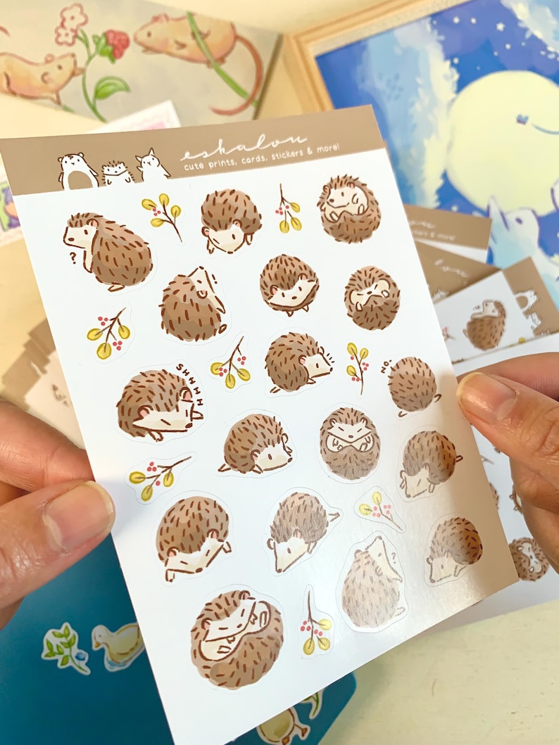 May include: A sheet of stickers featuring various illustrations of hedgehogs in different poses. The stickers are on a white background with a light brown border. The text "eshalou" and "cute prints, cards, stickers & more" is printed at the top of the sheet.