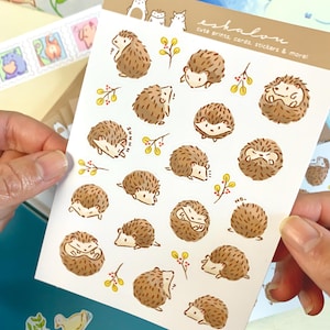 May include: A sheet of stickers featuring cute illustrations of hedgehogs in various poses. The stickers are on a white background with a light brown border. The text "eshalou cute prints, cards, stickers & more!" is printed in the top right corner.