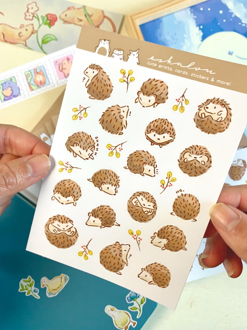 May include: A sheet of stickers featuring cute illustrations of hedgehogs in various poses. The stickers are on a white background with a light brown border. The text "eshalou cute prints, cards, stickers & more!" is printed in the top right corner.