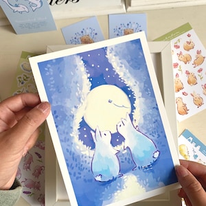 Op de afbeelding: Een ingelijste kunstdruk met twee blauwe konijnen die naar een volle maan kijken in een sterrennachtsc&egrave;ne. Rondom de print bevinden zich diverse stickers en kaarten met dierenillustraties, waaronder konijnen en beren.