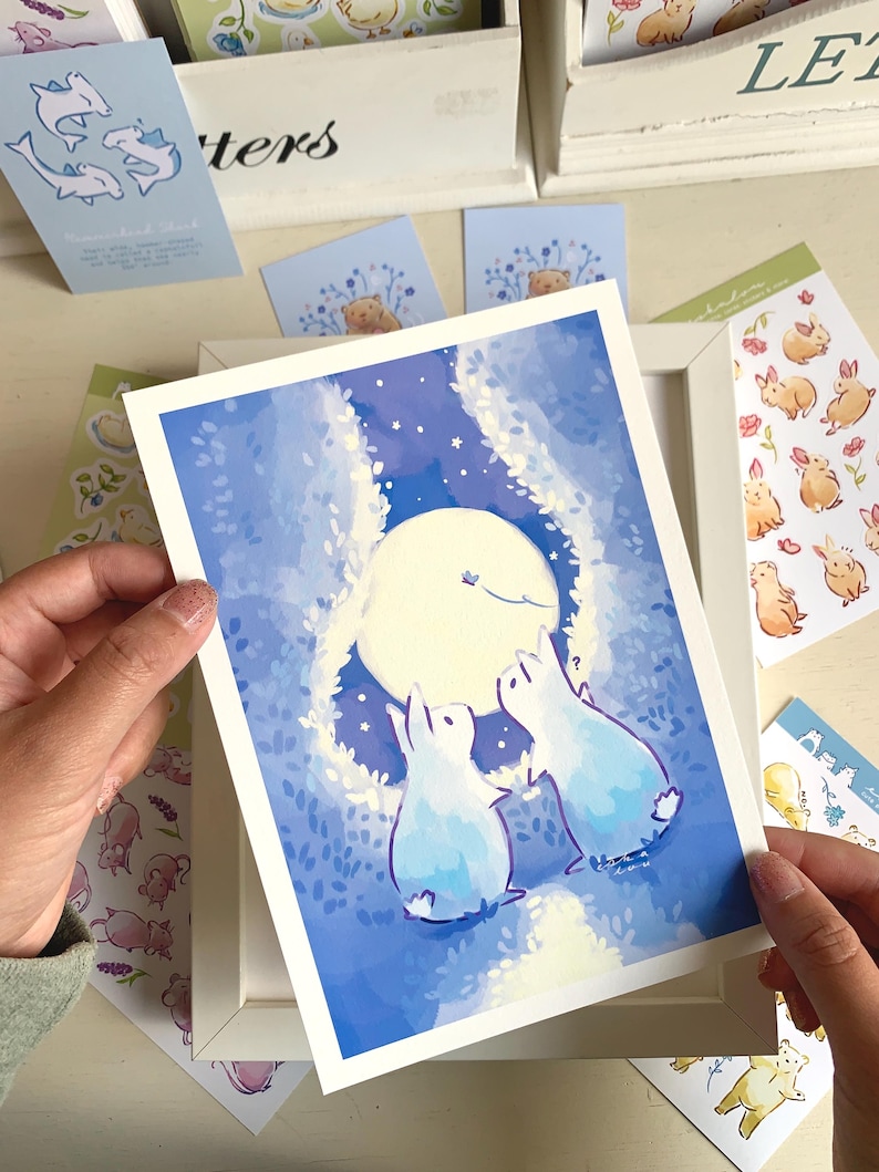 Op de afbeelding: Een ingelijste kunstdruk met twee blauwe konijnen die naar een volle maan kijken in een sterrennachtsc&egrave;ne. Rondom de print bevinden zich diverse stickers en kaarten met dierenillustraties, waaronder konijnen en beren.