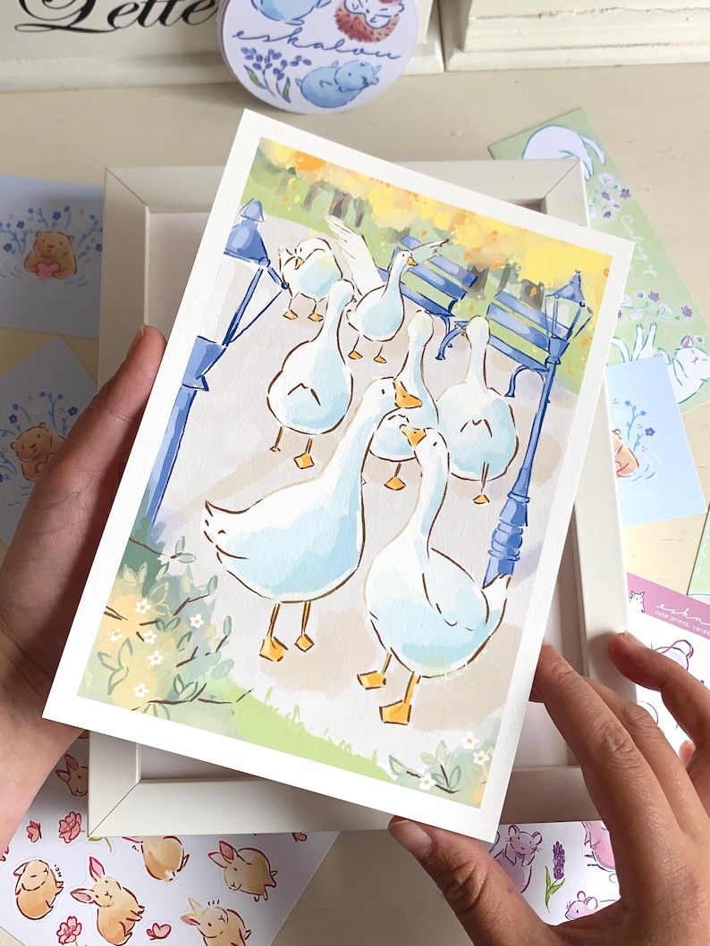 May include: Watercolor print of geese in a park setting, framed in white. The artwork features several white geese with yellow beaks and feet, walking on a path. Blue benches and lampposts are visible in the background, with a soft, pastel color palette.