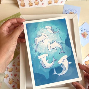 May include: A framed art print featuring cartoon hammerhead sharks swimming in a blue ocean. The artwork has a whimsical style with light blue and white sharks. Additional cards with bunny and bear illustrations are visible.
