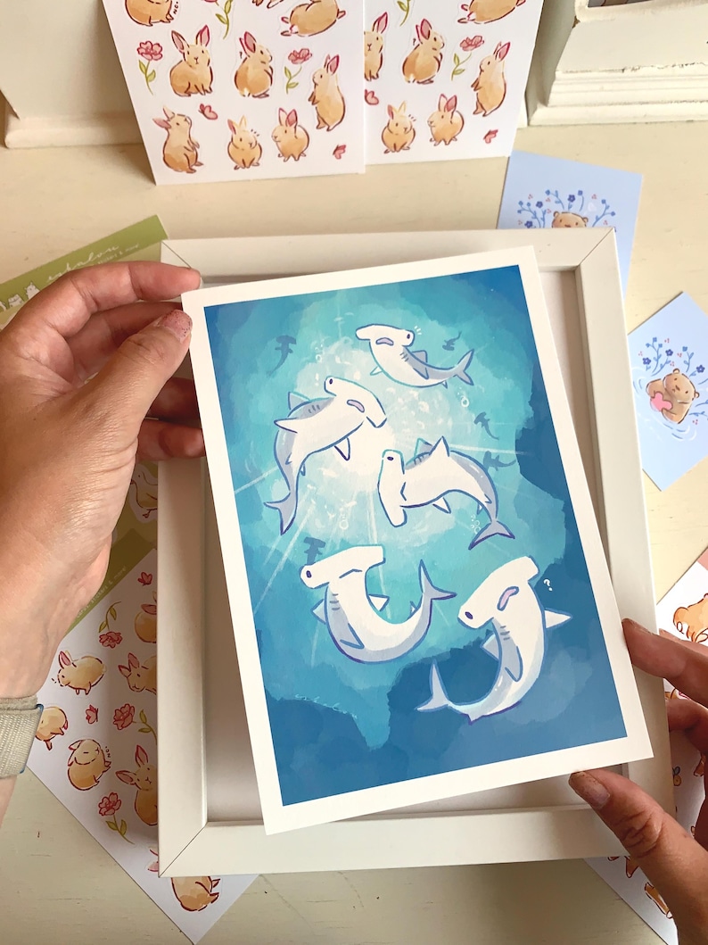 May include: A framed art print featuring cartoon hammerhead sharks swimming in a blue ocean. The artwork has a whimsical style with light blue and white sharks. Additional cards with bunny and bear illustrations are visible.