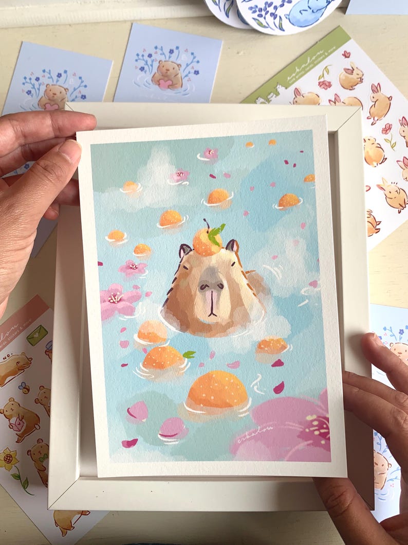 Op de afbeelding: Een ingelijste kunstdruk met een capibara die ontspant in een blauw zwembad met drijvende sinaasappels en roze bloemen. De capibara heeft een sinaasappel met een groen blad op zijn hoofd. Andere stickers en kaarten zijn zichtbaar.