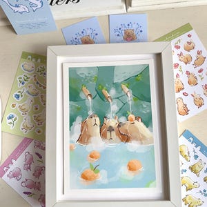 May include: A framed art print shows three capybaras in a hot spring with oranges. Various stickers with ducks, bears, and rabbits surround the print. The artwork is in a watercolour style with soft colours.