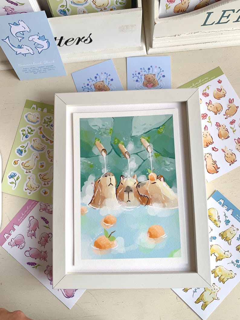 May include: A framed art print shows three capybaras in a hot spring with oranges. Various stickers with ducks, bears, and rabbits surround the print. The artwork is in a watercolour style with soft colours.