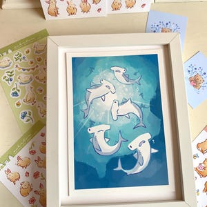 May include: A framed art print featuring cartoon hammerhead sharks swimming in a blue ocean. Surrounding the framed print are stickers and cards with illustrations of bunnies, ducks, and bears. The frame is white.