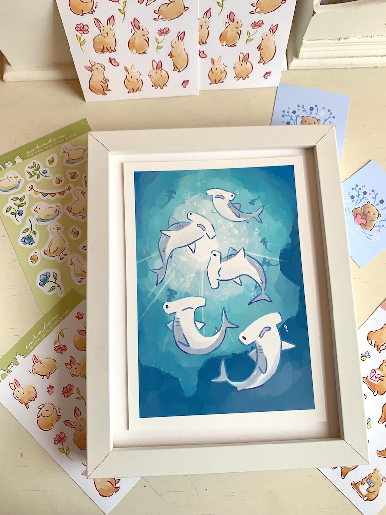 May include: A framed art print featuring cartoon hammerhead sharks swimming in a blue ocean. Surrounding the framed print are stickers and cards with illustrations of bunnies, ducks, and bears. The frame is white.