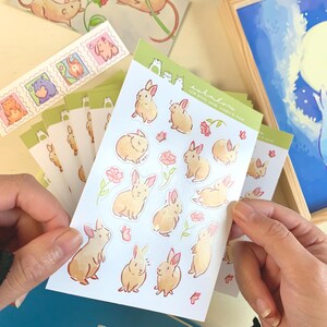 May include: A sheet of stickers featuring cute bunny illustrations with pink flowers and butterflies. The stickers are on a white background and have a green border. The text "toki no mori" is printed on the sheet, which translates to "forest of time".