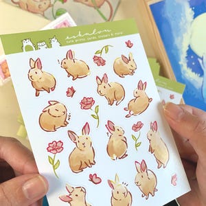 May include: A sheet of stickers featuring cute illustrations of brown rabbits with pink flowers and butterflies. The stickers are on a white background with the text "eshalon cute prints, cards, stickers & more".