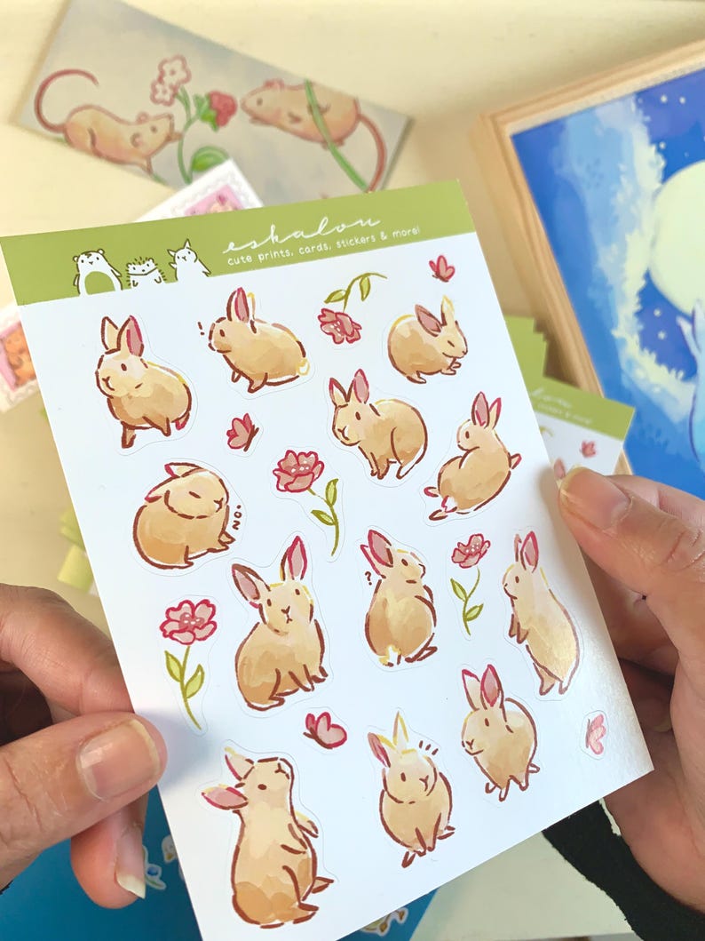 May include: A sheet of stickers featuring cute illustrations of brown rabbits with pink flowers and butterflies. The stickers are on a white background with the text "eshalon cute prints, cards, stickers & more".