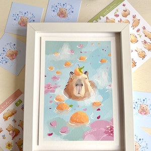 Op de afbeelding: Een ingelijste kunstdruk met een capibara die ontspant in een bad met sinaasappelfruit en roze bloemblaadjes. Stickers en kaarten met dier- en bloemillustraties omringen de witte lijst.