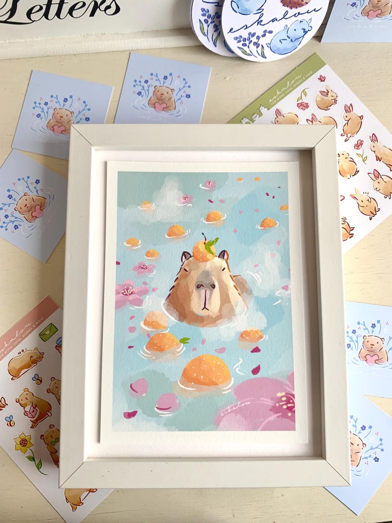 Op de afbeelding: Een ingelijste kunstdruk met een capibara die ontspant in een bad met sinaasappelfruit en roze bloemblaadjes. Stickers en kaarten met dier- en bloemillustraties omringen de witte lijst.