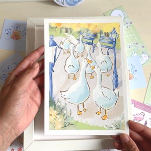 May include: A watercolor print featuring a group of white geese in a park setting. The artwork includes a blue bench, a lamppost, and yellow and green foliage. The print is held in a white frame, with additional stationery items visible.